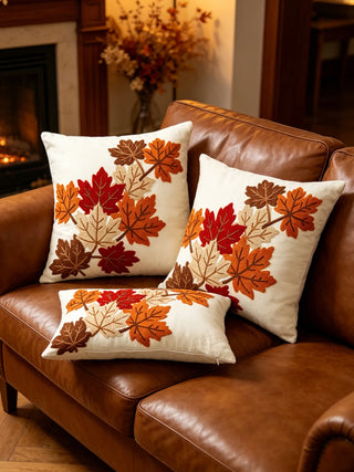 Three square throw pillows with embroidered autumn maple leaves in red, orange, brown, and beige, arranged on a brown leather sofa in a cozy living room setting.