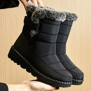 A pair of black mid-calf winter boots with gray faux-fur trim at the top, held up by hands against a light wood-paneled background.