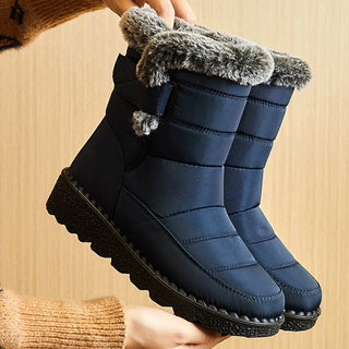 A pair of navy blue winter boots with gray faux-fur trim and a chunky black sole, being held up by hands wearing a brown knit sweater against a light wood background.